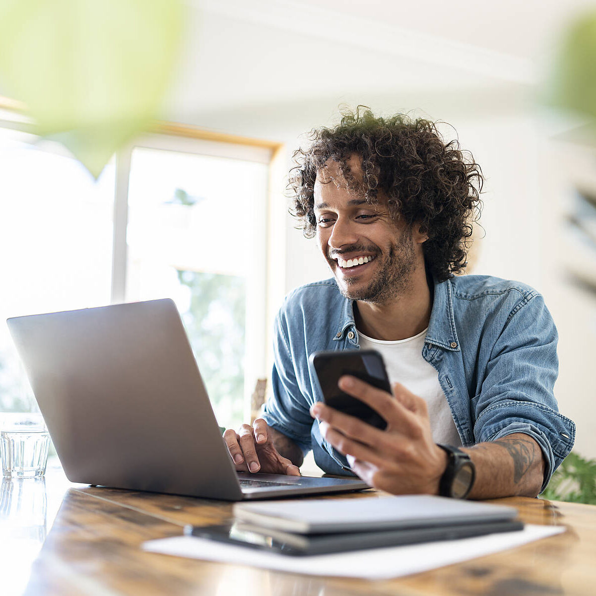 Handsome man at home, sitting at table looking at laptop and holding a smartphone